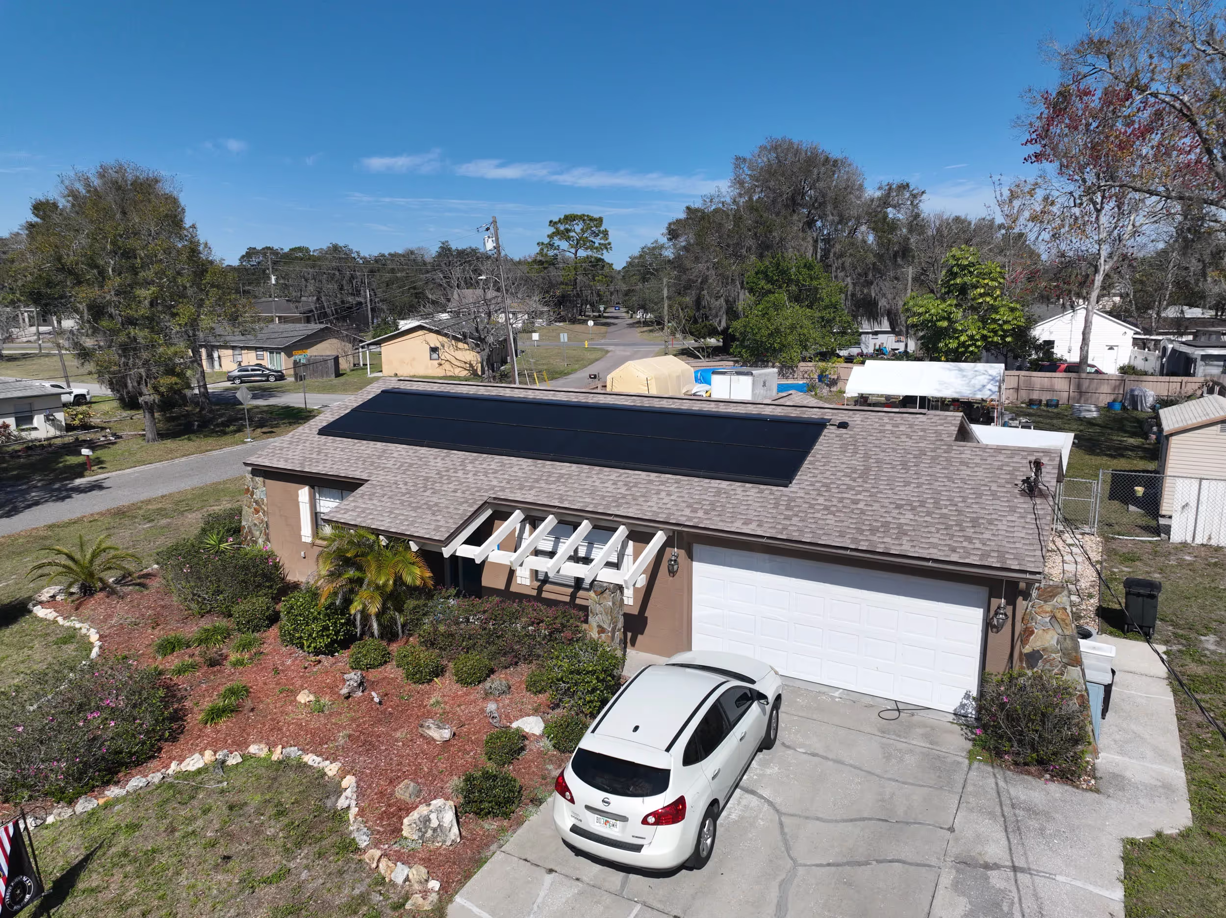 A single-story suburban house with solar panels on the roof, a white car parked on the driveway, and a landscaped front yard with shrubs and mulch.